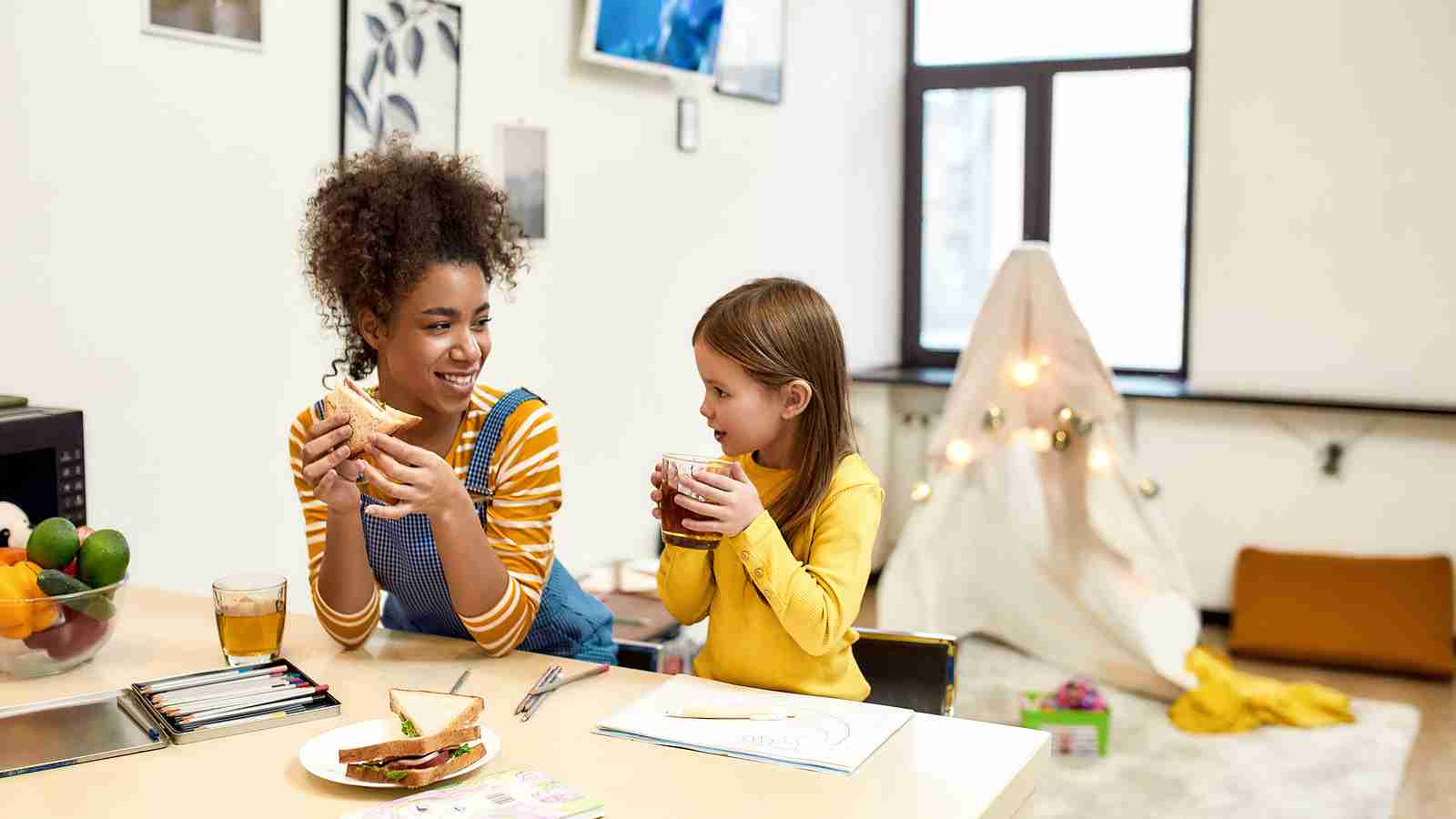 African american woman baby sitter entertaining caucasian cute little girl. Kid drinking tea while having lunch with her nanny. Leisure activities, babysitting concept. Horizontal shot